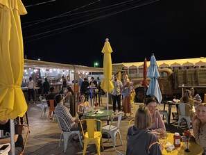 An outdoor dining area with tables and chairs, umbrellas, and a food stall in the background. - Mountain View Bungalows Under the Stars-Unit B Venus (Joshua Tree)