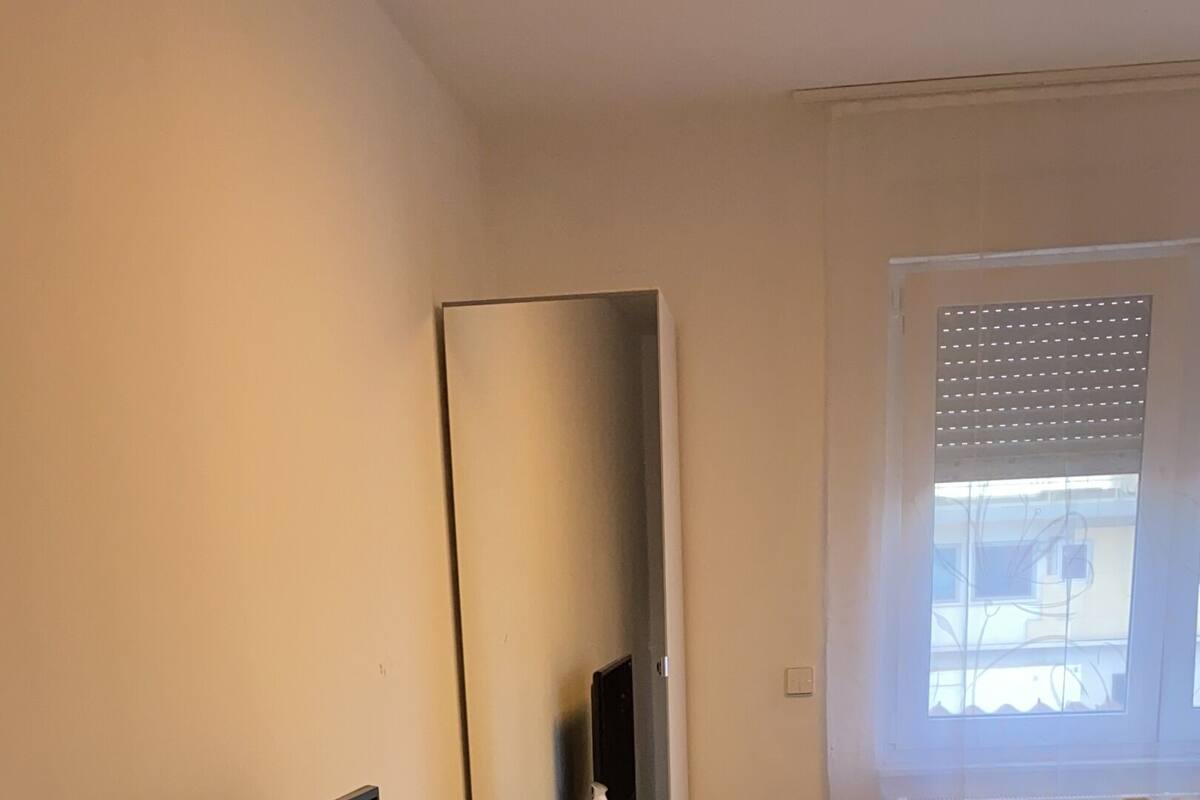 A modern bedroom with a bed, a desk with a computer, a mirror, and a window with blinds.