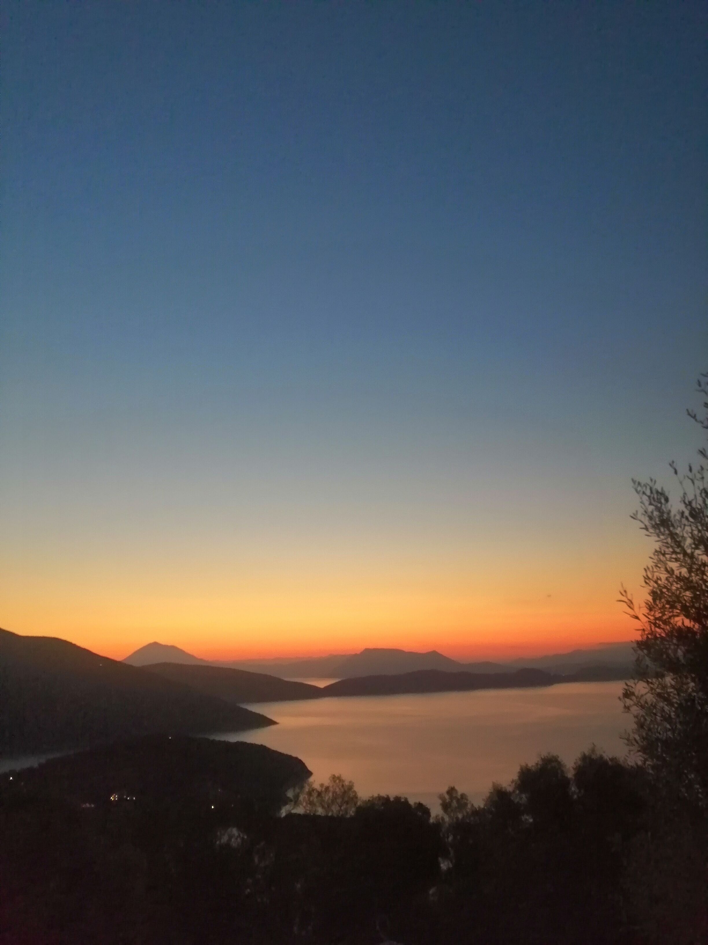 Une vue panoramique d’un paysage côtier au coucher du soleil, avec une mer calme et des montagnes lointaines.