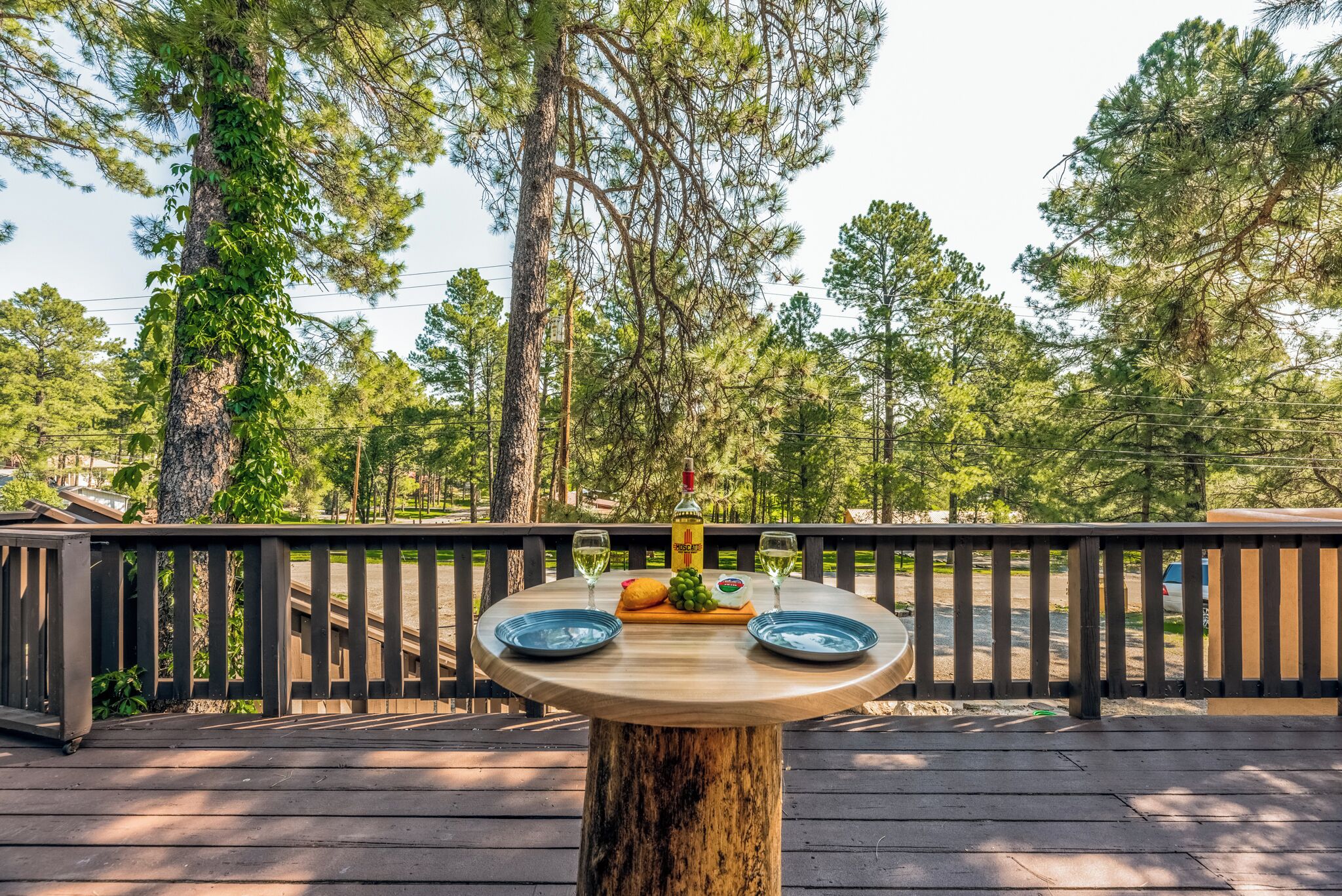 Een houten terras met een ronde tafel gedekt voor twee personen, inclusief een fles wijn, een brood en een schaal fruit.