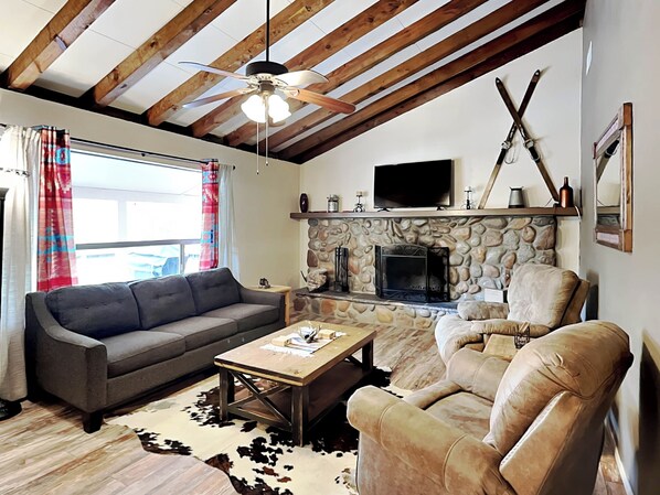 A living room with a stone fireplace, a television mounted above it, and a ceiling fan. - Elks Landing by Ruidoso Vacation Rentals (Ruidoso)