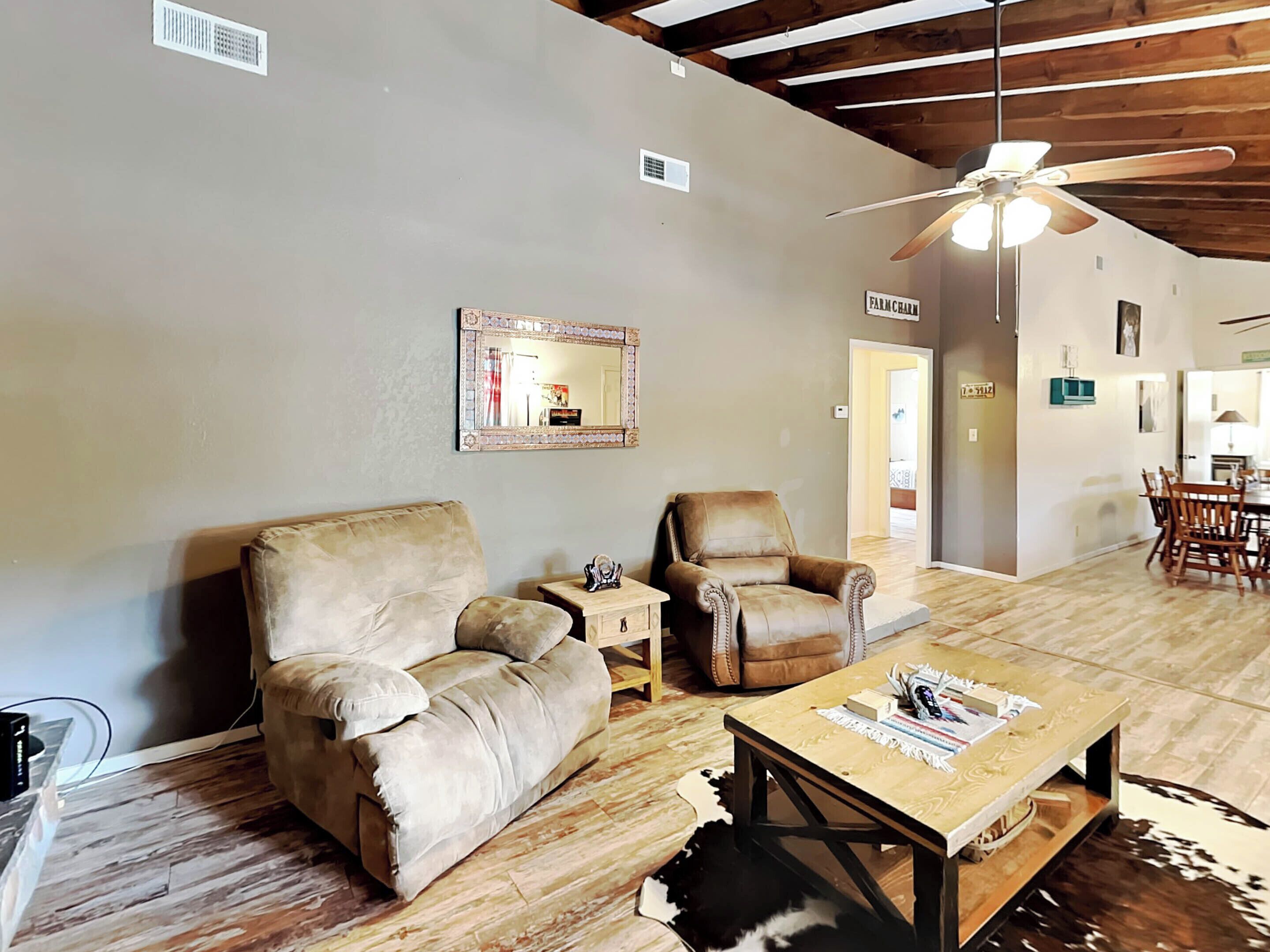 A spacious living room with a high ceiling, a ceiling fan, a large mirror, a leather armchair, a wooden coffee table, and a dining area in the background.