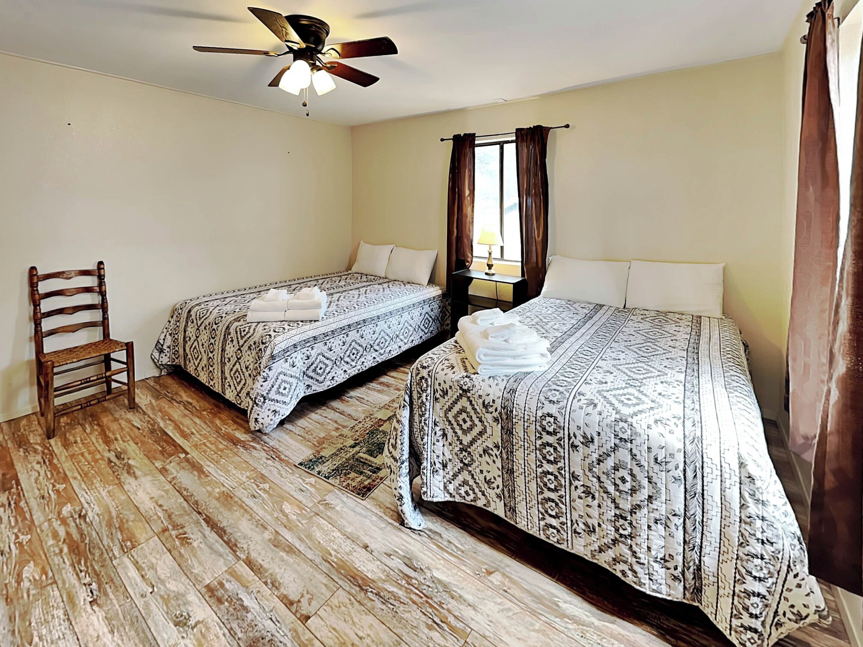 A room with two beds, a wooden chair, a ceiling fan, and a window with curtains.
