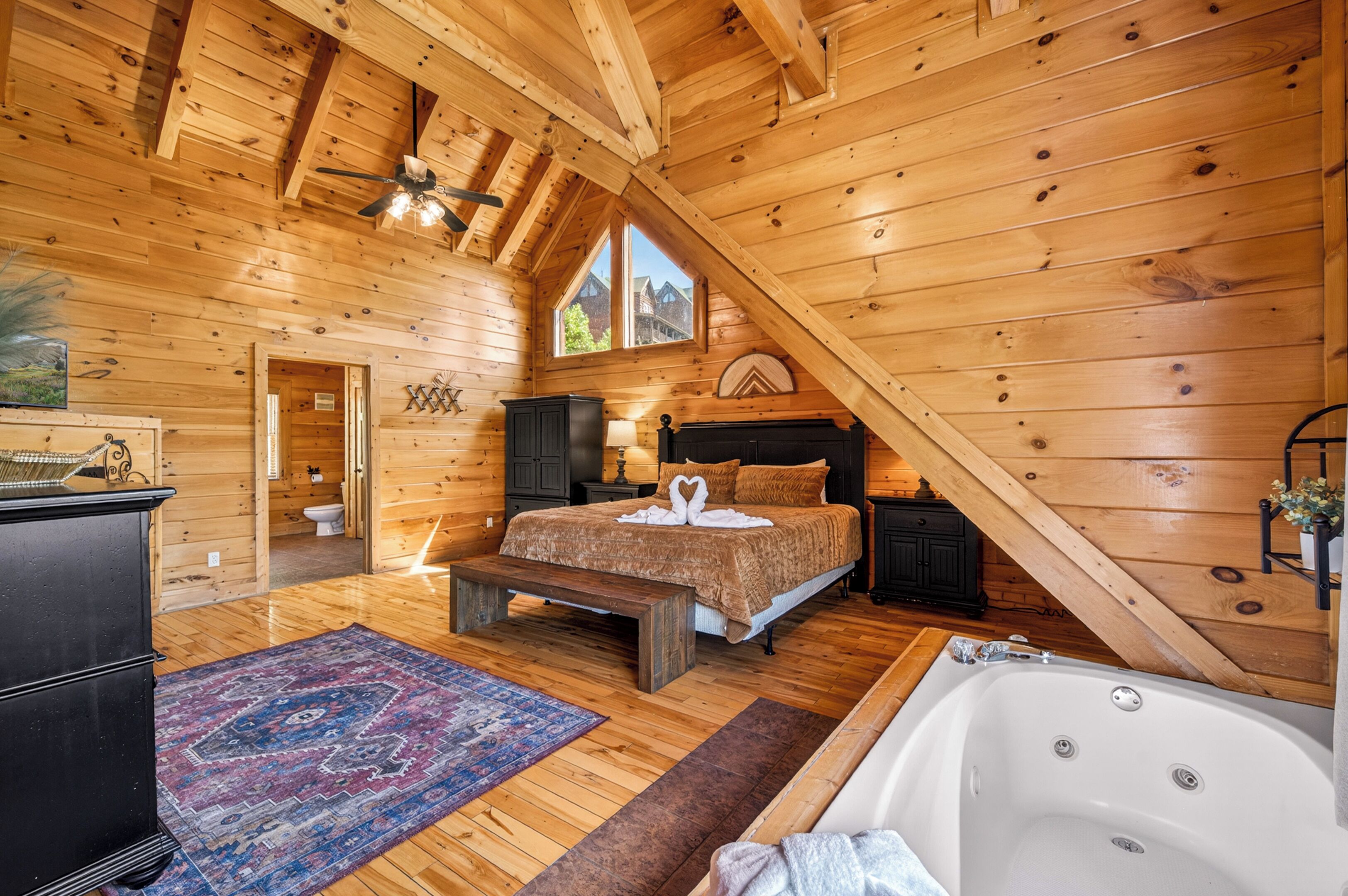 Une chambre située dans un chalet en rondins de bois, avec un plafond en bois, un grand lit, un banc, une baignoire et une vue sur l’extérieur.