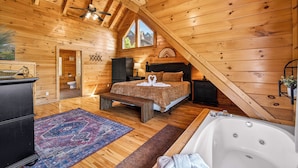A log cabin bedroom with a wooden ceiling, a large bed, a bench, a bathtub, and a view of the outside. - Timberwolf Vista w HotTub Pool View (Sevierville)
