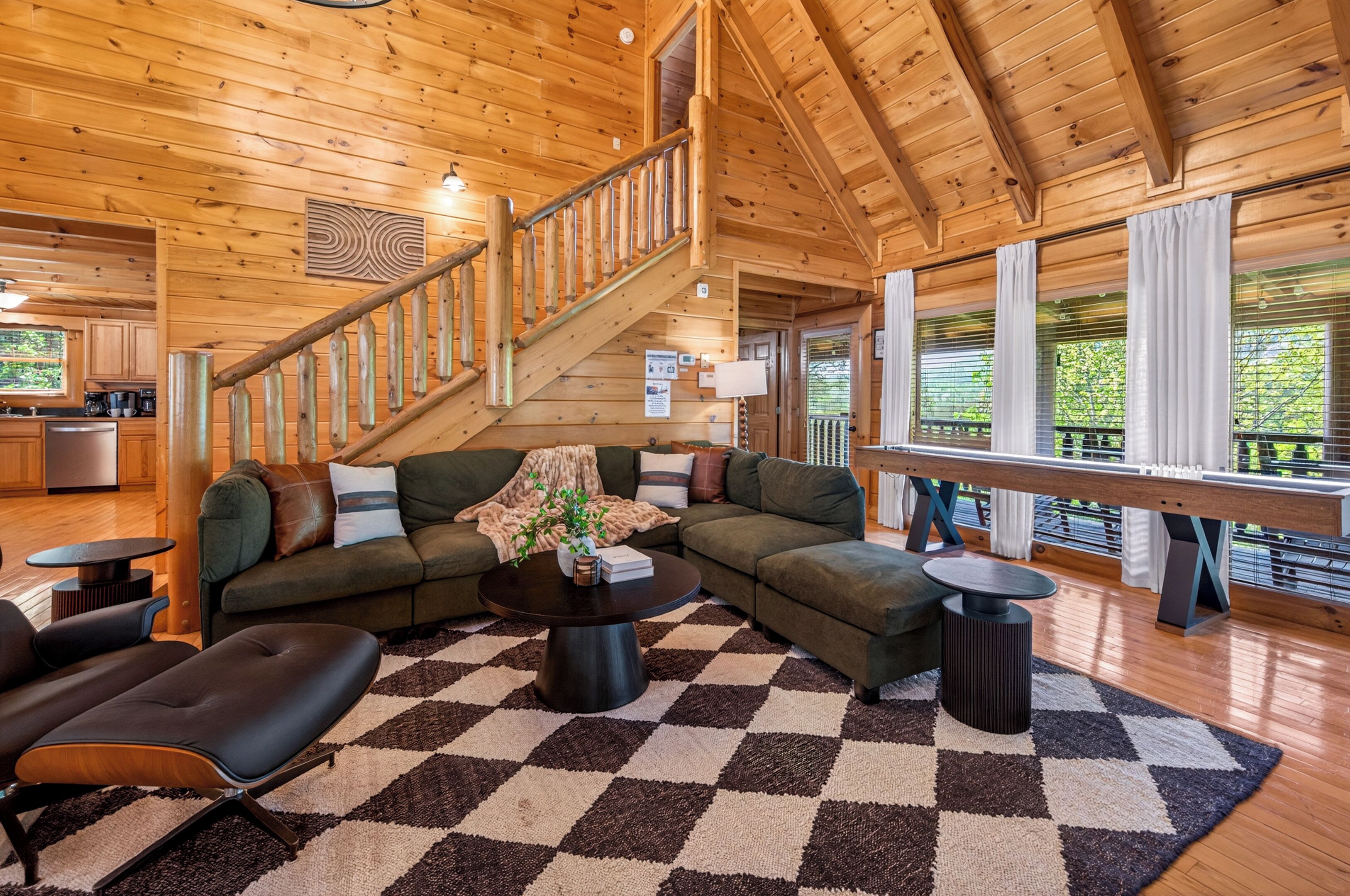 A wooden interior with a staircase, a sectional sofa, a coffee table, and a large window with curtains.