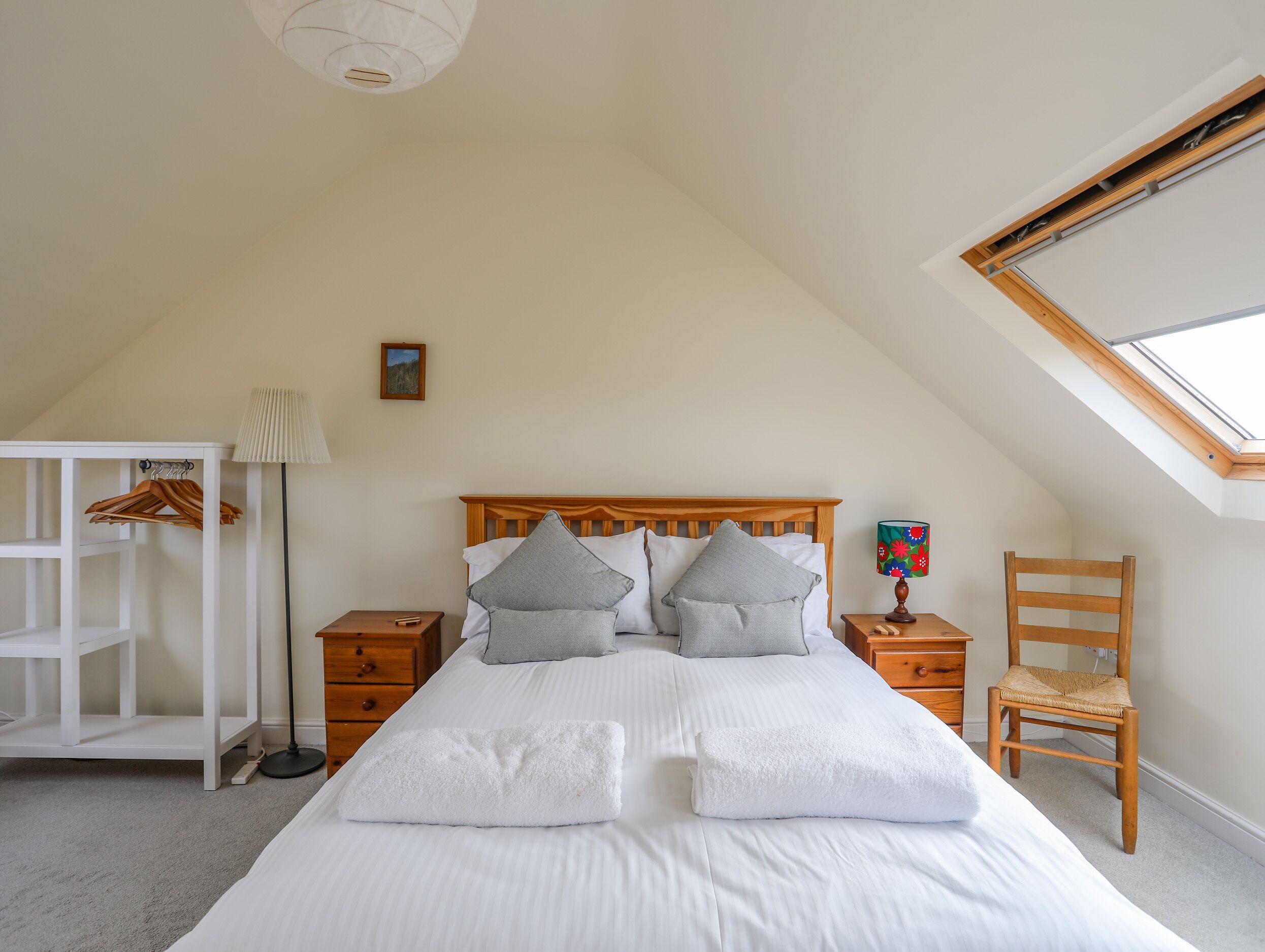 Ein Schlafzimmer mit schräg steilendem Dach, einem Holzbettgestell, zwei Nachttischen, einem Sessel, einem Kleiderschrank und einem Dachfenster.
