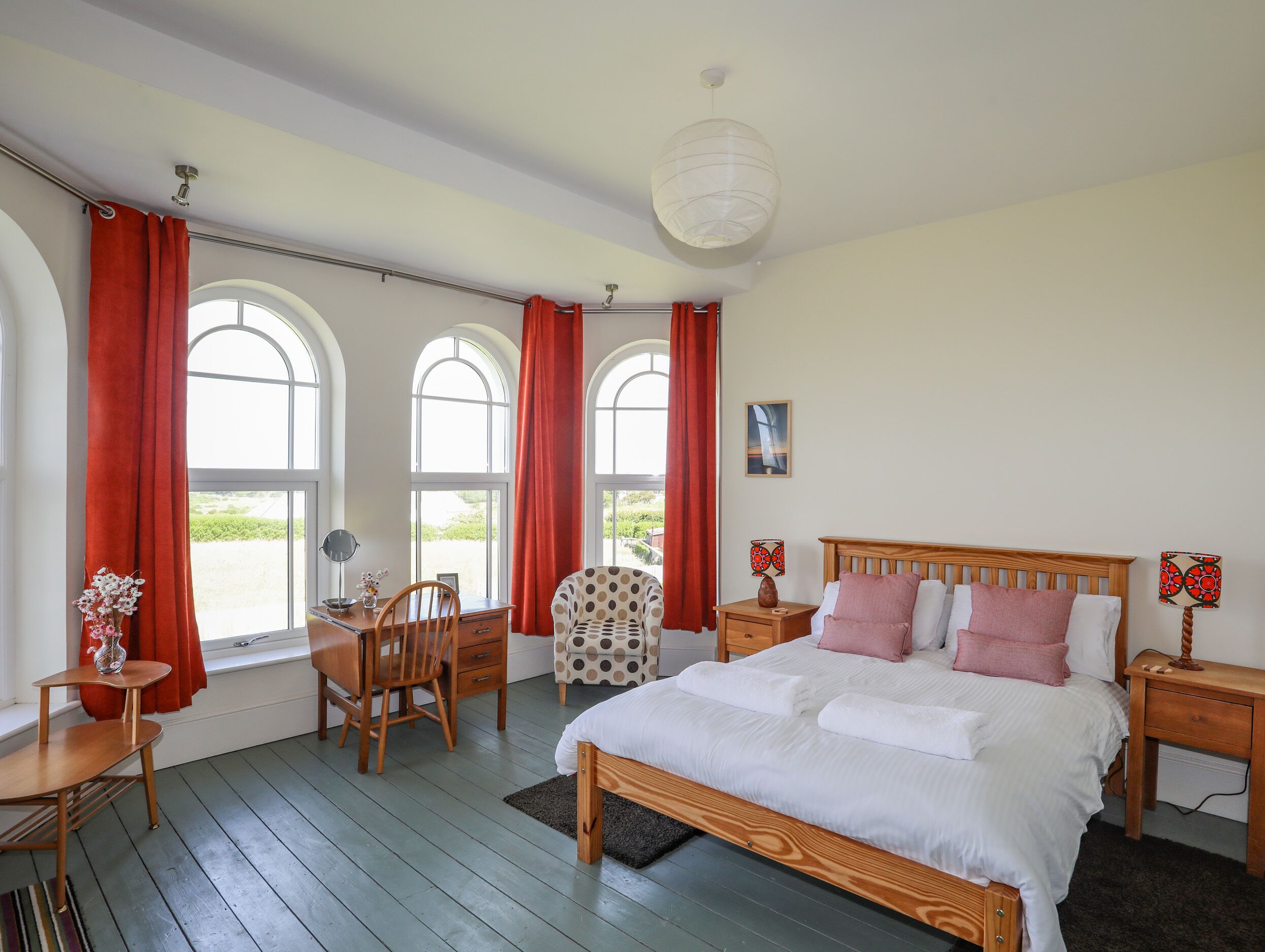A bedroom with a wooden bed, red curtains, a desk with a chair, and a view through arched windows.