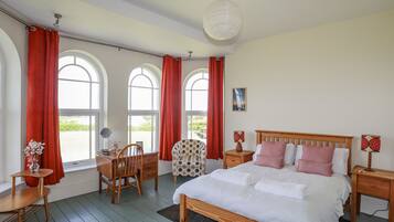 A bedroom with a wooden bed, red curtains, a desk with a chair, and a view through arched windows.
