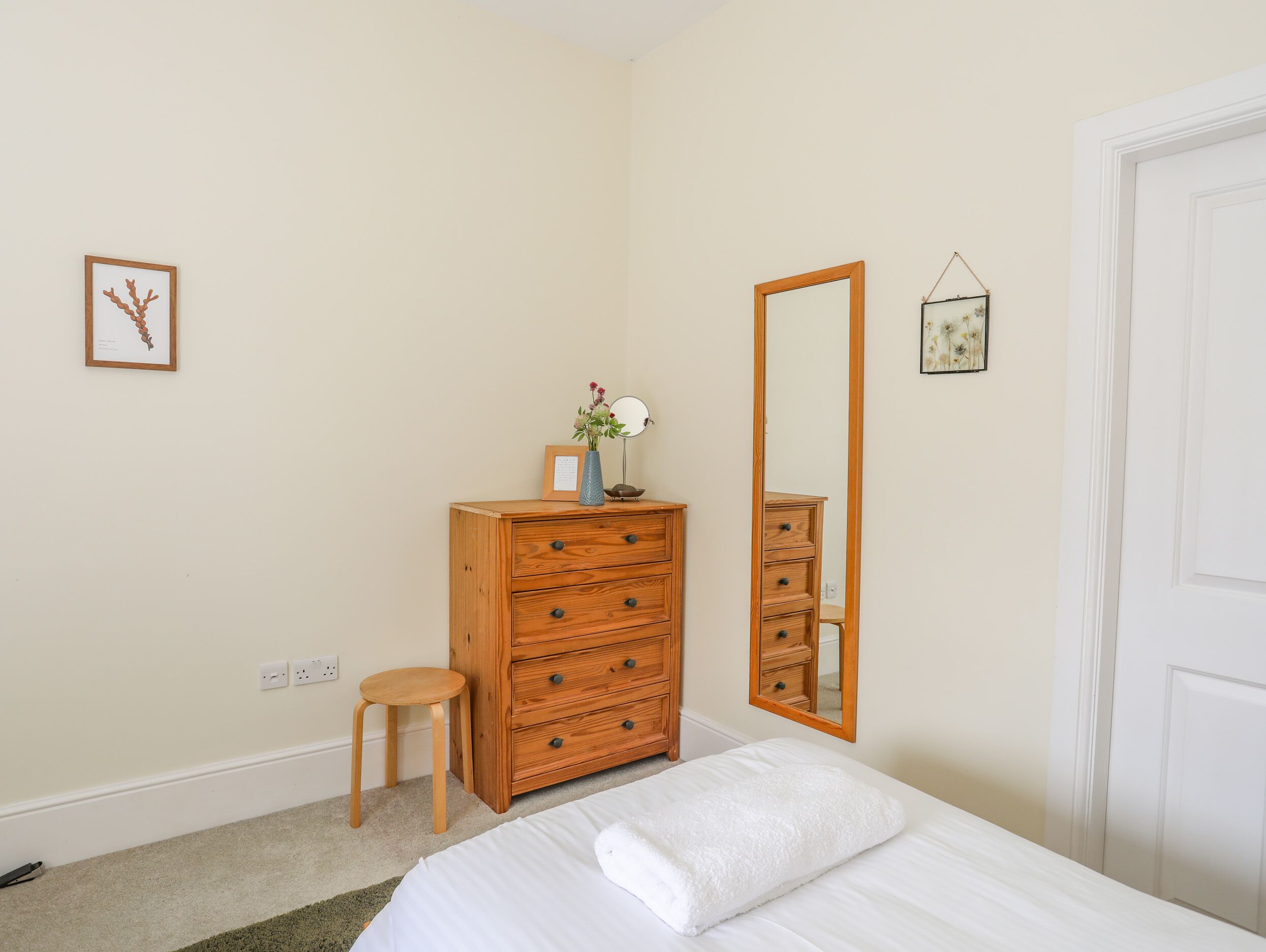 A bedroom with a wooden dresser, a mirror, a small stool, and a bed with a white comforter.