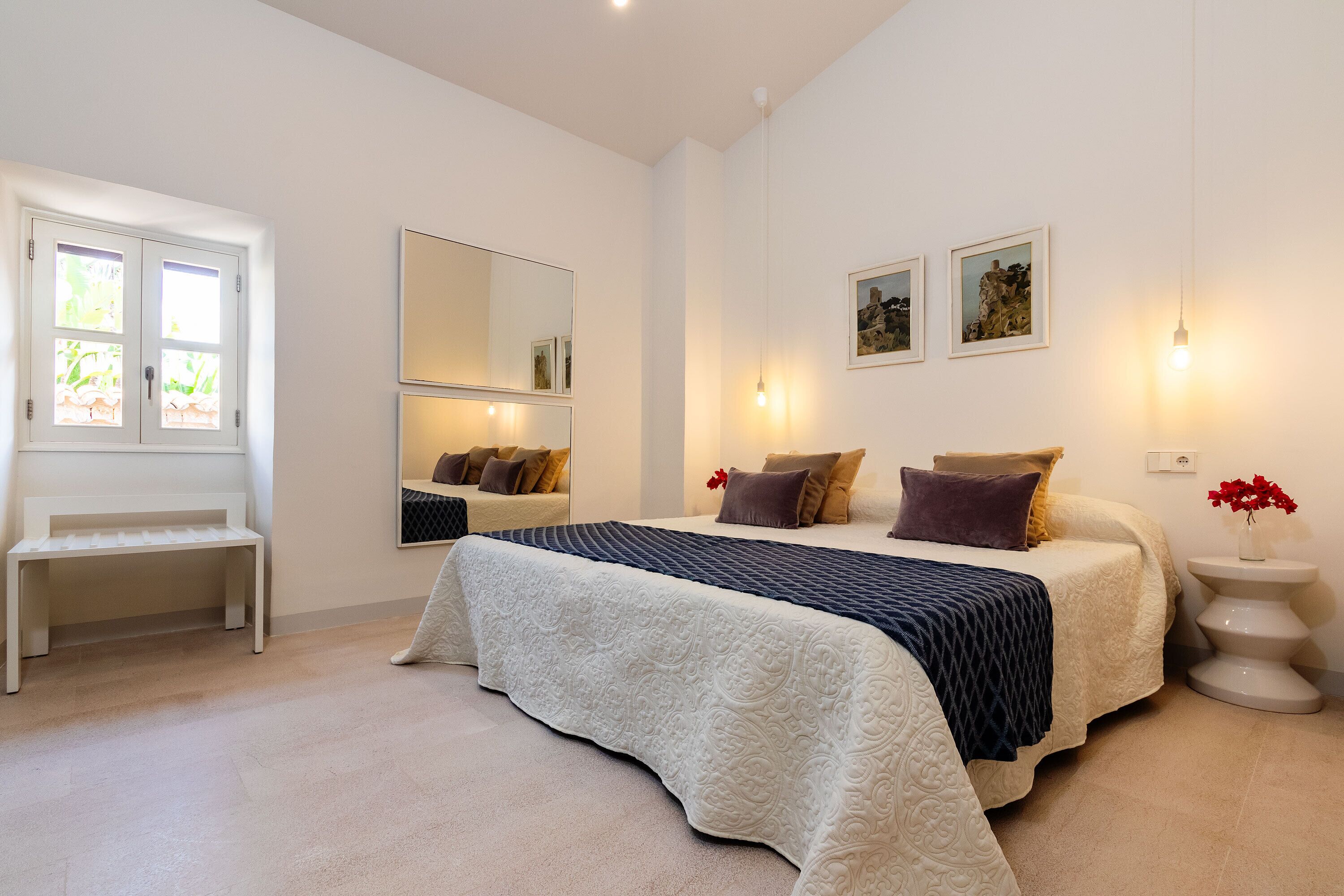 A bedroom with a bed, a bench, a mirror, and artwork on the wall.