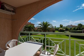 Ein Balkon mit weißem Tisch und Stühlen, einem grünen Geländer und Blick auf eine gepflegte Rasenfläche und entfernte Gebäude.