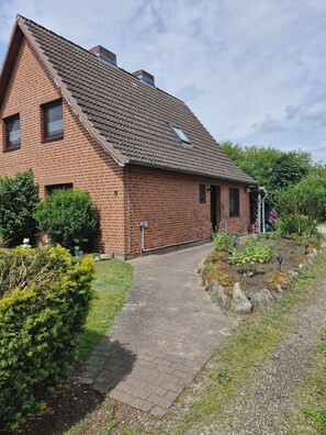 A brick house with a sloped roof, a paved driveway, and a garden with various plants and a small pond.