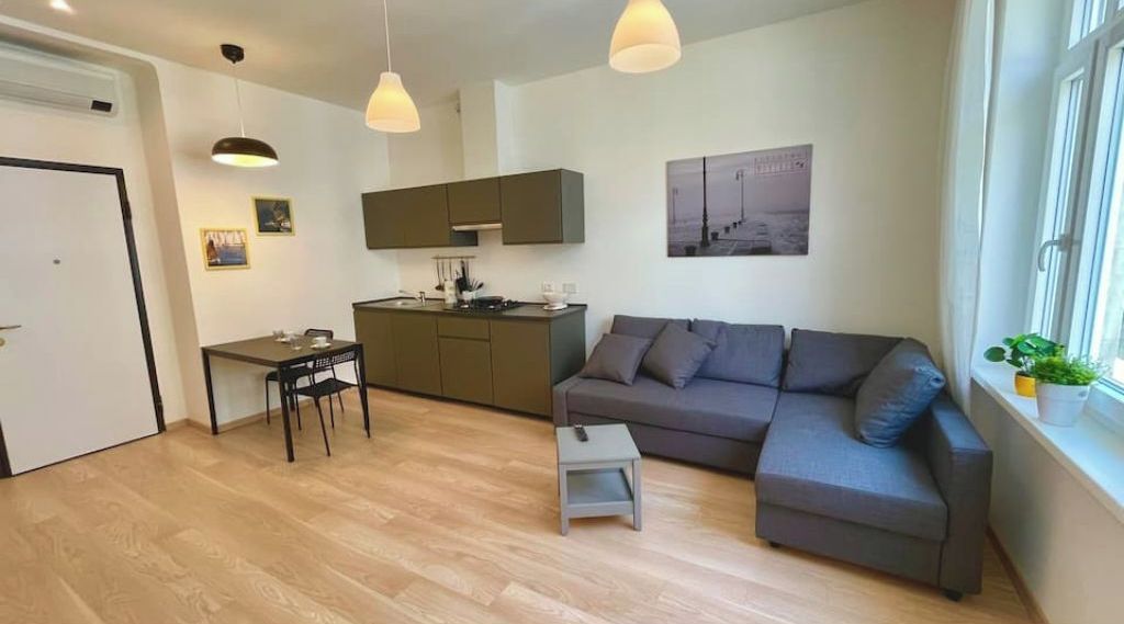 Un salon moderne avec un canapé gris, une petite table et un coin cuisine comprenant des placards et une table à manger.