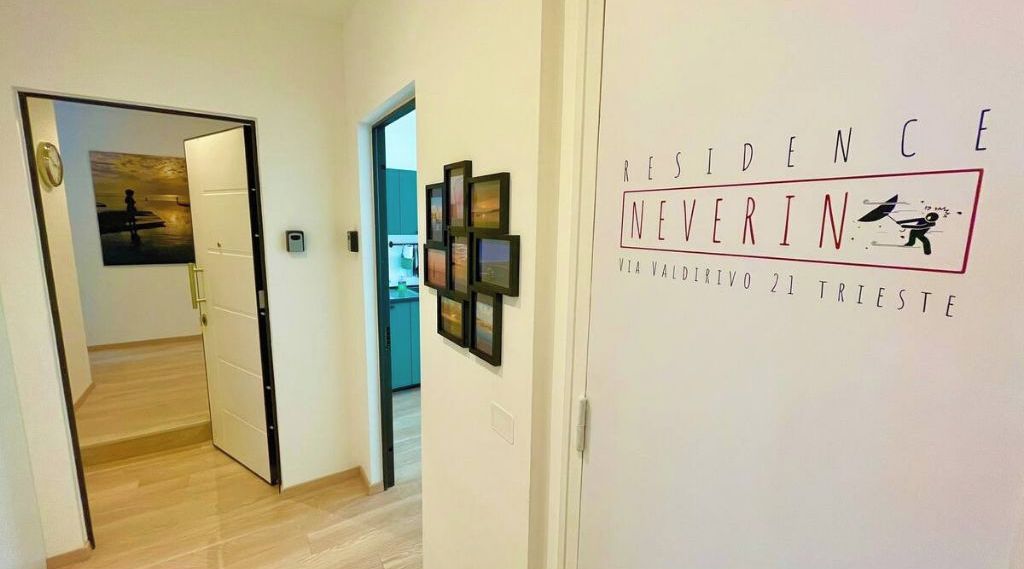 A hallway with a door leading to a room, a wall with framed pictures, and a sign that reads "RESIDENCE NOVERIN VIA VALDERIVO 21 TRIESTE".