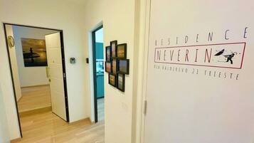 A hallway with a door leading to a room, a wall with framed pictures, and a sign that reads "RESIDENCE NOVERIN VIA VALDERIVO 21 TRIESTE".
