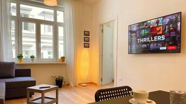 Ein modernes Wohnzimmer mit einer Couch, einem kleinen Tisch, einem Fernseher, der einen Thriller zeigt, und einem Fenster mit Vorhängen.