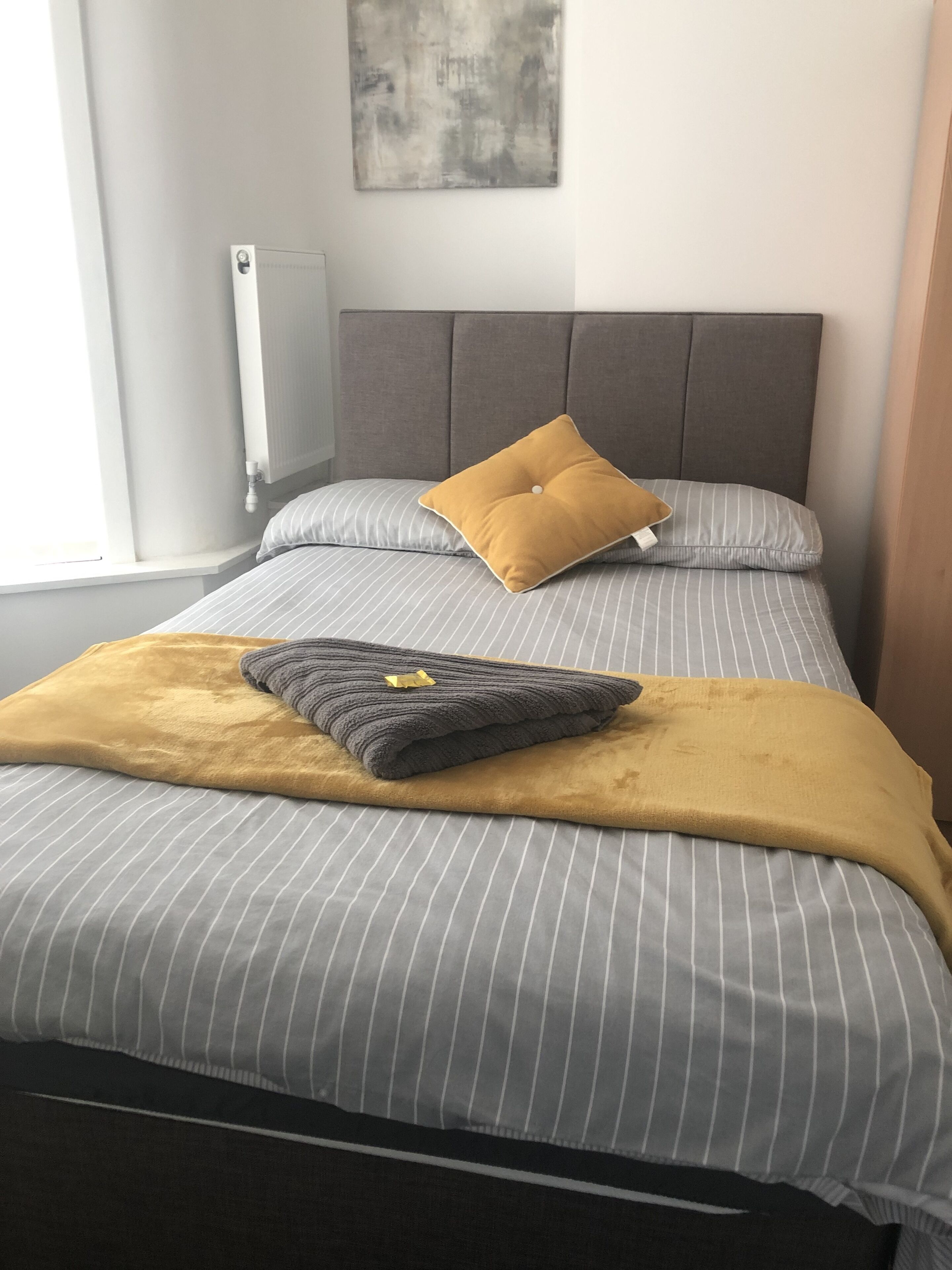 Een net opgemaakt bed met gestreept beddengoed, een mosterdkleurige plaid en twee kussens, waarvan één mosterdkleurig is.