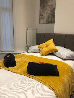 A neatly made bed with a yellow throw blanket, a black cap, and a black pillow on a white and gray striped bedspread.