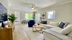 Amplia sala de estar con un sofá blanco, un sillón azul, una mesa de centro de madera y un televisor grande. Hay plantas en macetas y un ventilador de techo.