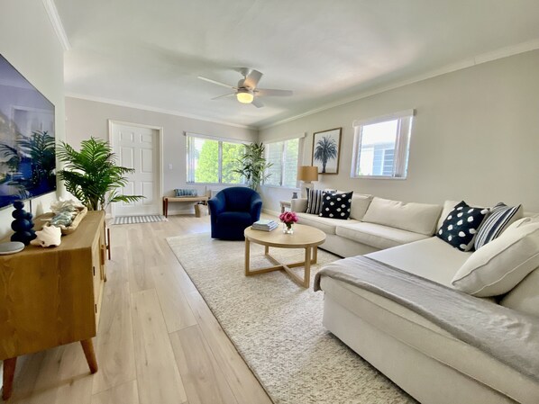 A spacious living room with a white sofa, a blue armchair, a wooden coffee table, and a large TV. There are potted plants and a ceiling fan.