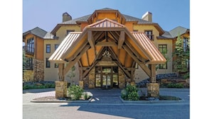 Un grand bâtiment à plusieurs étages, doté d’une structure en bois remarquable à l’avant, qui pourrait servir d’entrée, de réception ou d’espace d’accueil, et entouré d’un aménagement paysager soigné et d’une allée pavée.