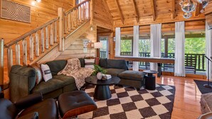 A wooden cabin interior with a staircase, a living area with a checkered rug, and large windows. - Lux Lodge! Pool-Sauna-HotTub-Game-Pet Frndly (Sevierville)