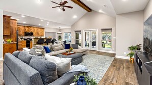 A spacious living area with a large gray sectional sofa, a wooden kitchen with built-in cabinets, a ceiling fan, and a large TV on a black stand.