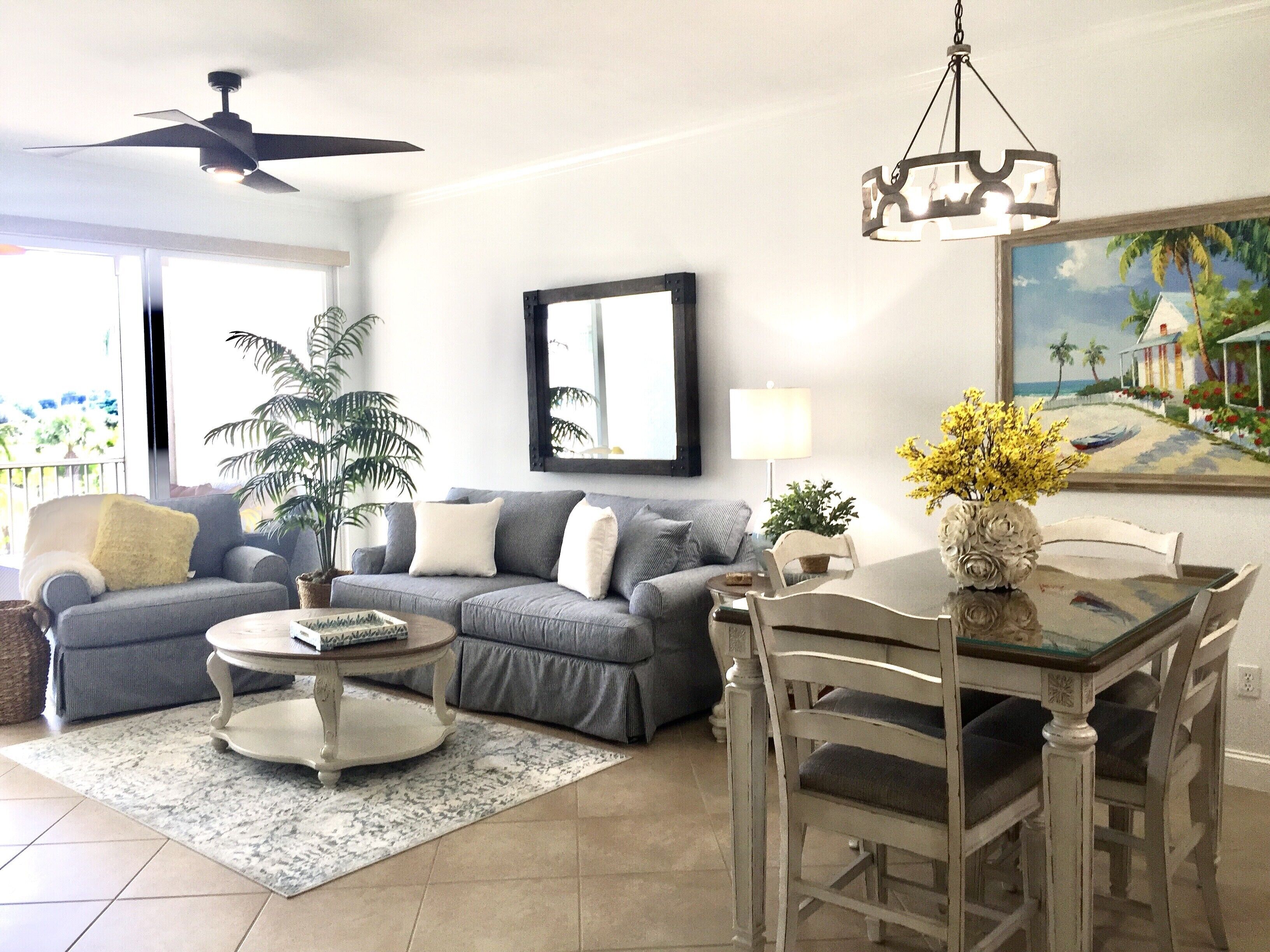 A living room with a grey sofa, a round coffee table, a dining table with chairs, a ceiling fan, and a painting of a beach scene.