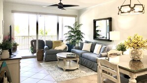 Ein geräumiges Wohnzimmer mit einer großen Couch, einem Couchtisch und einem Esstisch. Es gibt einen Deckenventilator, ein großes Fenster und einen Blumentopf.