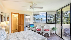 Un dormitorio con cama, televisión, dos sillones de mimbre con cojines rojos, ventilador de techo y balcón con vistas al mar y a los árboles.