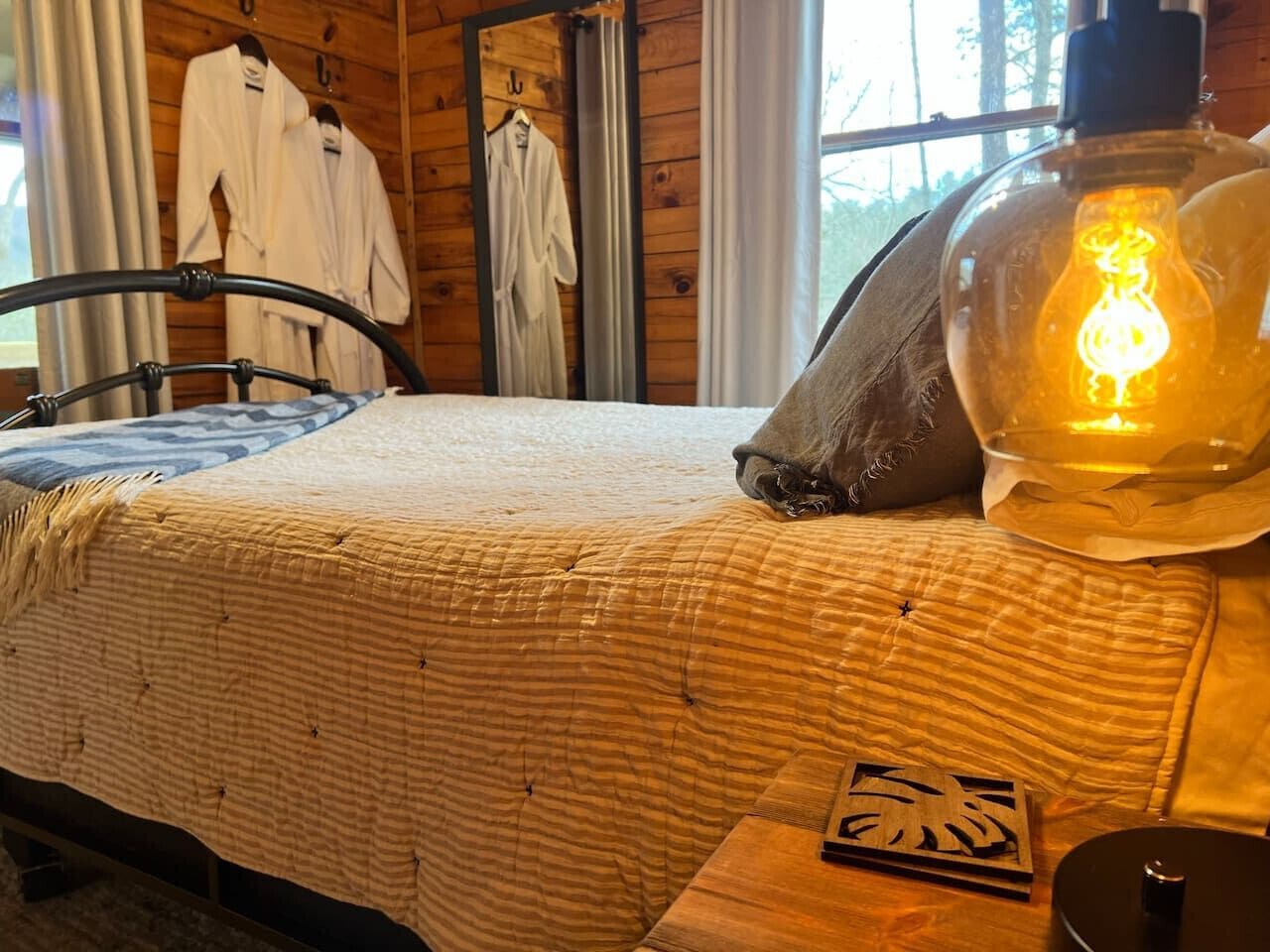 A bed with a quilted blanket, a wooden headboard, a bedside table with a lamp, and a window with curtains.