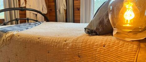 A bed with a quilted blanket, a wooden headboard, a bedside table with a lamp, and a window with curtains.