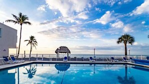 A clear blue pool with lounge chairs and palm trees, surrounded by a fence and a building.