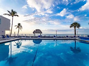 A clear blue pool with lounge chairs and palm trees, surrounded by a fence and a building.