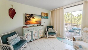 A room with a white tiled floor, a white wicker sofa, a green armchair, a wooden cabinet, and a painting of a sunset.
