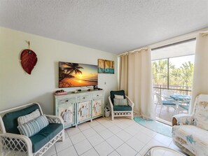 A room with a white tiled floor, a white wicker sofa, a green armchair, a wooden cabinet, and a painting of a sunset.