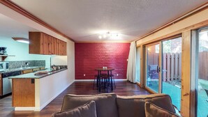 Eine Küche mit Holzschränken, einer Mikrowelle und einer Kaffeemaschine. Es gibt einen Essbereich mit Tisch und Stühlen. Der Raum verfügt über eine rote Backsteinwand und eine Glastür, die in einen Hinterhof führt.