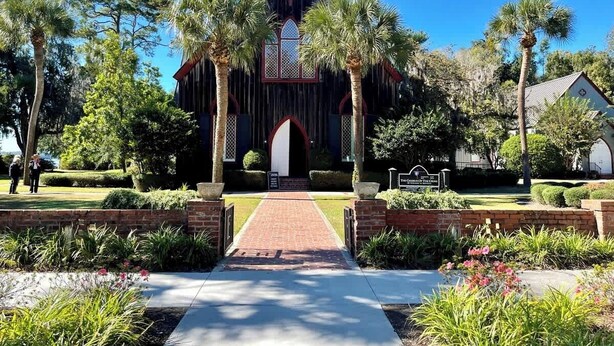 A church with a central arched entrance, surrounded by palm trees and a brick pathway leading to the door.