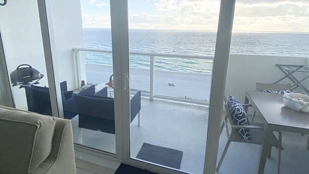 Una habitación con un amplio ventanal corredera que da a un balcón con vistas al océano. Hay un sofá y una pequeña mesa con una silla en el balcón.