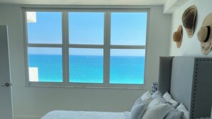 Un dormitorio con un ventanal grande con vistas al mar, una cama con ropa blanca y una mesita de noche con una lámpara y un objeto decorativo.