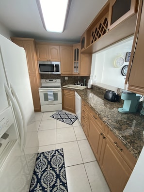 Compact kitchen with modern design, wooden cabinetry, and granite countertop, featuring a microwave, oven, coffee maker, sink, and small rug.