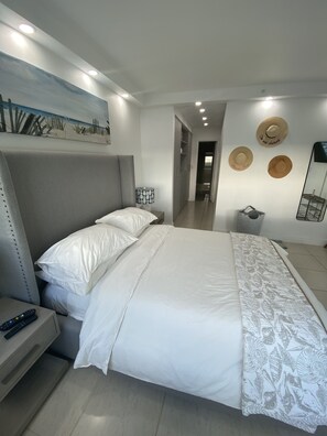Relax in a stylish room with a comfortable bed, bedside table, and lamp, surrounded by a beach-themed painting and decorative hats.