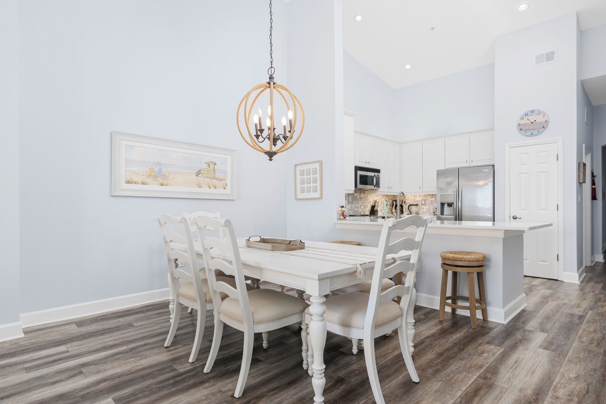 A dining area with a white table and chairs, a chandelier, and a kitchen in the background.