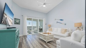 A spacious living room with a white couch, a coffee table, and a teal cabinet. There is a ceiling fan, a TV mounted on the wall, and a view of the balcony through the glass door.