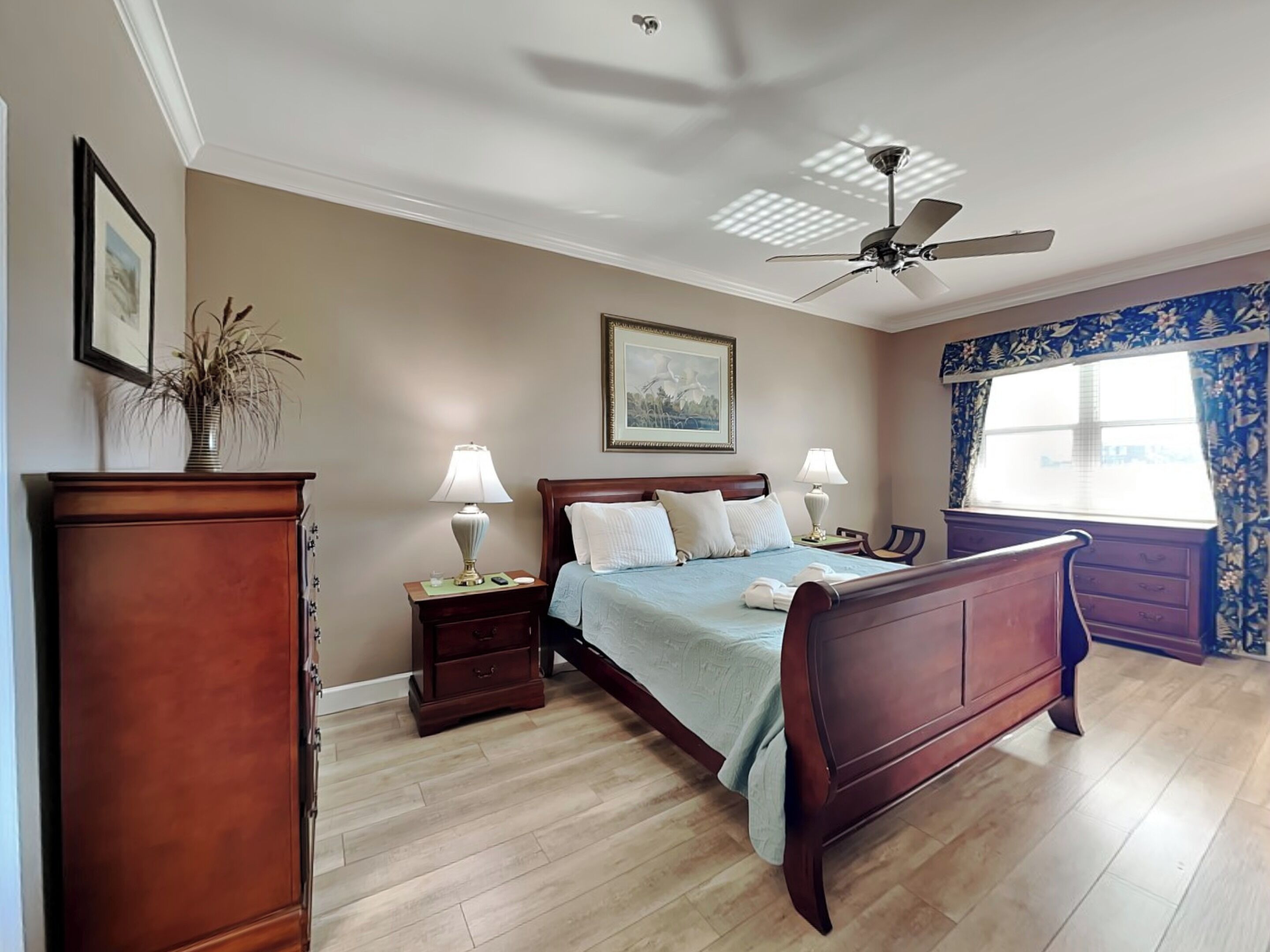 A bedroom with a wooden bed, nightstands, a ceiling fan, a window with curtains, and a painting on the wall.