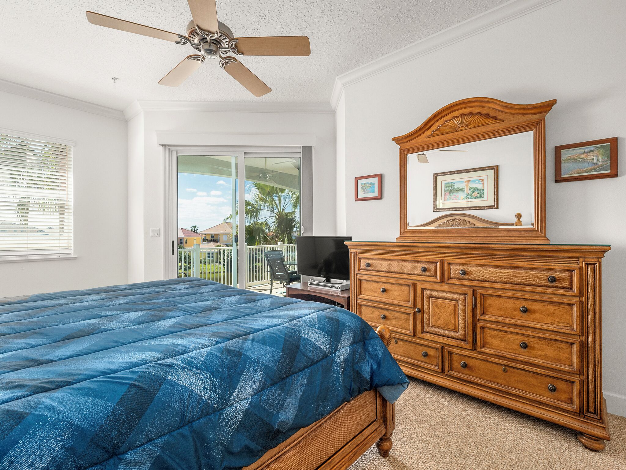 A bedroom with a bed, a dresser, a ceiling fan, and a balcony with a view of palm trees.