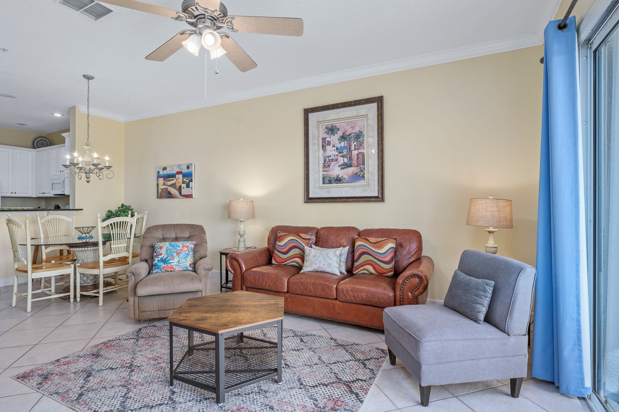 A living room with a brown leather sofa, a grey armchair, a wooden coffee table, and a patterned rug. There is a ceiling fan, a chandelier, and a framed painting on the wall.