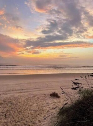 A beach with a sunset sky, sandy shore, and some vegetation in the foreground.