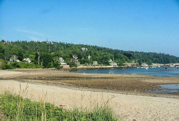 Une plage avec des maisons construites sur une colline, un rivage sablonneux et des bateaux sur l’eau.